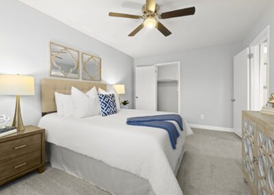 A cozy bedroom featuring a ceiling fan above a neatly made bed, side table with books and lamp