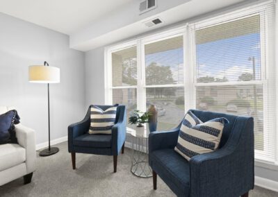 A cozy living room featuring two blue chairs, lamp, corner table with vase and a white couch